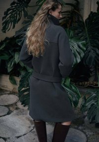 Lungo cappotto grigio con collo alto, vita aderente e maniche lunghe. Abbinato a stivali marroni alti fino al ginocchio, con sfondo di fogliame verde.