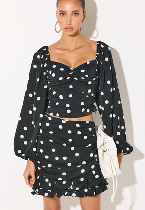 Femme portant un crop top à pois noirs à manches longues et une jupe à volants assortie, tenant un sac blanc avec cordon, faisant face.