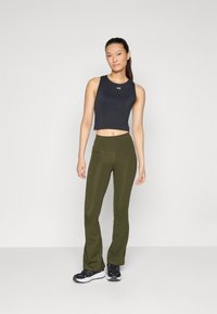 Schwarzes, ärmelloses, kurzes Oberteil und olivgrüne, ausgestellte Yogahosen, beide aus dehnbarem Material. Schwarz-weiße Sportschuhe.