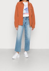 Veste en velours côtelé amples de couleur orange brûlé, associée à un t-shirt graphique blanc et à un jean taille haute bleu clair. Baskets blanches.