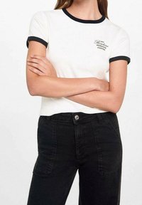 T-shirt bianca con colletto e maniche a coste nere, con la scritta "LA MIA RUTINA DEL CAFFÈ MATTUTINO", abbinata a jeans neri a vita alta.