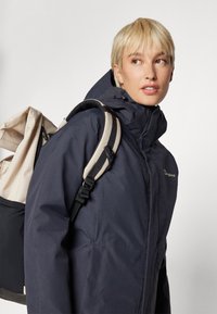 Navy blue waterproof jacket with a high collar, front zipper, and adjustable hood; paired with a beige backpack featuring patterned accents.
