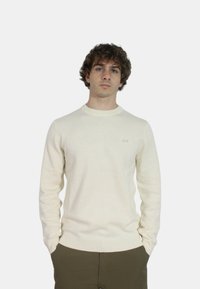 Maglione lavorato a maglia color crema con collo a girocollo a coste, maniche lunghe e dettaglio logo sul petto. Indossato sopra pantaloni verde oliva.
