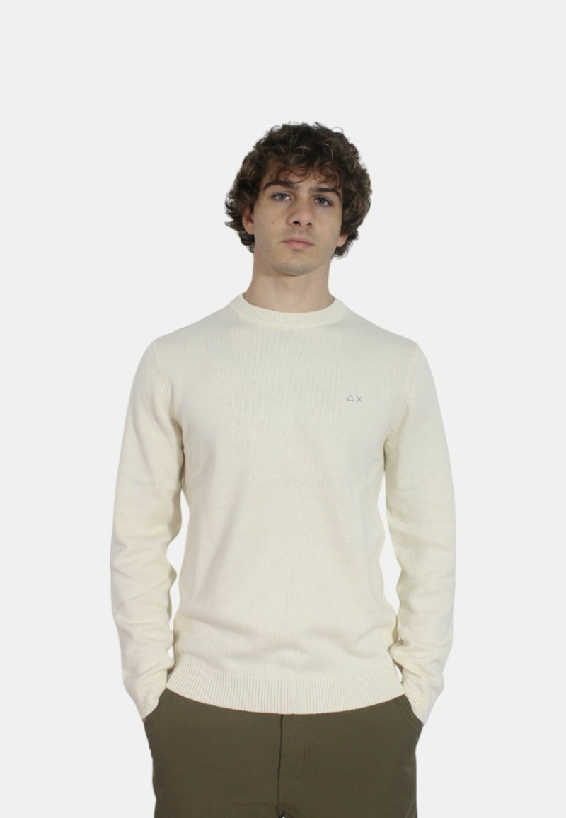 Maglione lavorato a maglia color crema con collo a girocollo a coste, maniche lunghe e dettaglio logo sul petto. Indossato sopra pantaloni verde oliva.