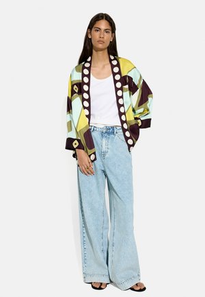 Kobieta w luźnych jasnoniebieskich dżinsach, białym tank topie, wielokolorowej kurtce w stylu kimono i czarnych butach z odkrytymi palcami, stojąca na jednolitym tle.