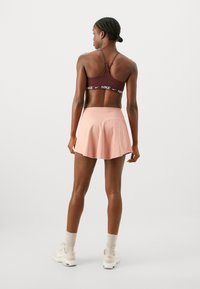 Sujetador deportivo burdeos con logo blanco, combinado con una falda acampanada de color rosa claro. Material suave y estilo casual, complementado por zapatillas blancas.