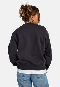 Felpa nera con colletto e polsini a coste, realizzata in tessuto morbido. Indossata sopra una camicia grigia, abbinata a jeans blu chiaro.