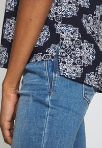 Granatowa bluzka w białe i niebieskie kwiatowe wzory, z bocznym rozcięciem, w połączeniu z jasnoniebieskimi dżinsami. Gładka tekstura, luźny krój.