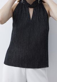 Žena v černém bezramenném halter topu s jemnými metalickými pruhy a bílých kalhotách si upravuje vlasy na jednobarevném bílém pozadí.