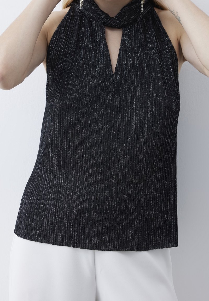 Žena v černém bezramenném halter topu s jemnými metalickými pruhy a bílých kalhotách si upravuje vlasy na jednobarevném bílém pozadí.