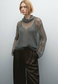 Gray, loose-knit sweater with a cowl neckline, layered over a brown high-waisted pant. Soft texture, semi-sheer design, and relaxed fit.