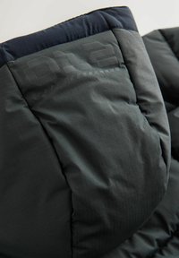 Gros plan sur la manche d'une veste matelassée foncée avec un motif alphanumérique embossé et une bordure bleu marine au poignet.