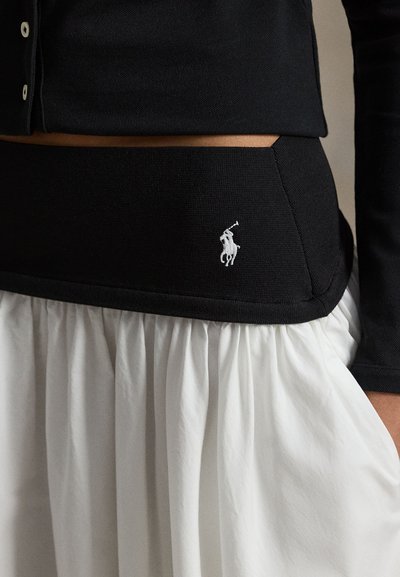 Haut noir ajusté avec une finition texturée, associé à une jupe plissée blanche. Comprend un petit logo blanc brodé sur la ceinture.