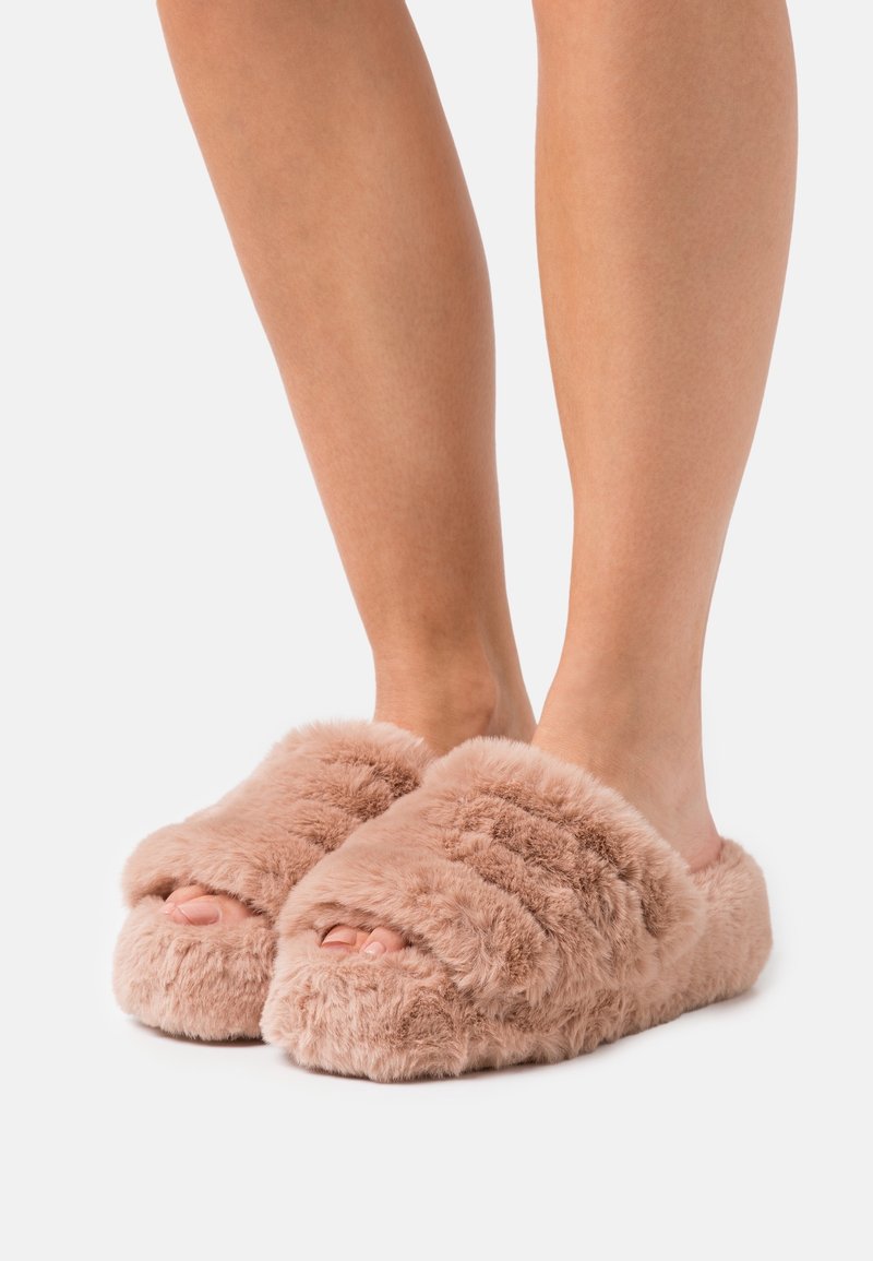 Pieds portant des chaussons ouverts du bout des orteils, duveteux et rose pâle, avec une texture épaisse et douce sur un fond clair uni.