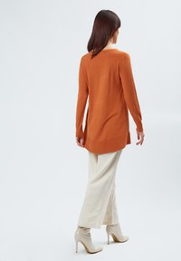 Langarm, verbrande-oranje trui met een iets gebogen zoom, gecombineerd met crème-wijd been broek en beige stilettohakken.