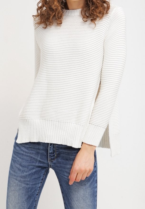 Femme portant un pull en maille côtelée blanche avec des fentes sur les côtés, associée à un jean en denim bleu, debout devant un fond clair uni.