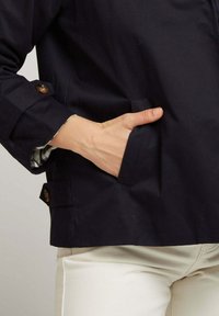Veste navy avec une poche frontale, des accents de boutons sur les manches et une doublure à carreaux visible aux poignets.