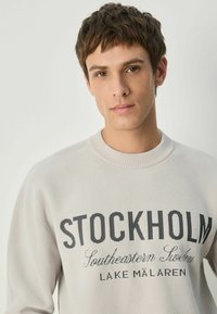 Lys grå sweater med "STOCKHOLM" i fede sorte bogstaver, med ribbet krave og blød tekstur, mod en neutral baggrund.