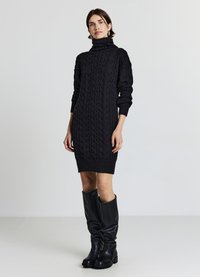 Vestito felpa nero lavorato a maglia con collo alto, polsini a costine e silhouette aderente. Abbinato a stivali neri in pelle morbida che arrivano al ginocchio.