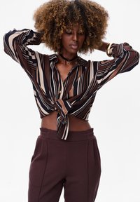 Striped button-up shirt in brown, cream, and navy, tied at the waist. Paired with high-waisted, dark wine-colored pants with pockets.