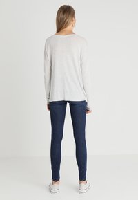 Maglietta grigia chiara a maniche lunghe con taglio dritto, abbinata a jeans skinny blu scuro e sneakers bianche in tela vista da un angolo posteriore.