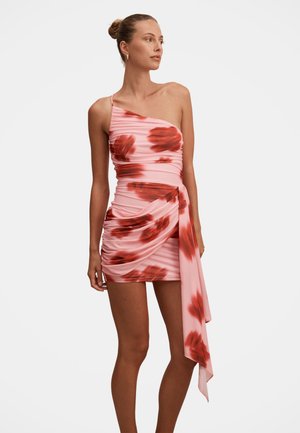 Γυναίκα που φοράει ένα ροζ μίνι φόρεμα με έναν ώμο, διακοσμημένο με κόκκινα λουλουδένια σχέδια και με μια μακριά draped ταινία στη μία πλευρά, στέκεται μπροστά σε λευκό φόντο.