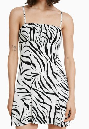 Nainen, jolla on yllään hihaton, mustavalkoinen seeprakuosinen minimekko ohuilla olkaimilla, kuvattuna hartioista reisiin asti.