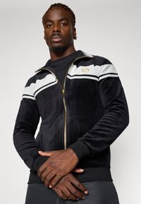 Svart zip-up jakke i mykt stoff, med hvite og grå striper, gull-logo og ribbede mansjetter og hem. Modellen poserer med armene krysset.