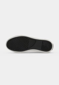 Semelle de chaussure en caoutchouc noir avec des motifs géométriques et le nom de la marque "Guess" embossé au centre, bord blanc autour de la semelle.