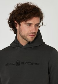 Sudadera con capucha de color gris oscuro que cuenta con un cordón en la capucha, tejido texturizado y un logotipo bordado de "SAIL RACING" en el frente.
