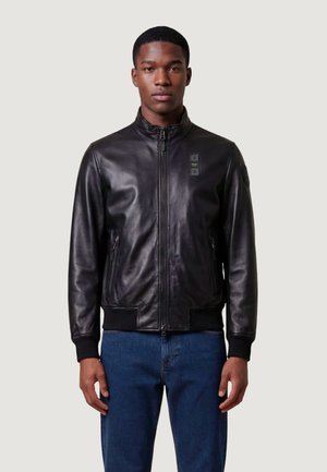 Młody mężczyzna w czarnej skórzanej kurtce z kieszeniami na suwak i niebieskich dżinsach, stojący na jednolitym jasnym tle.