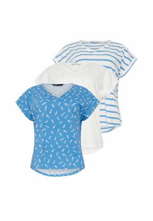 Frau mit lockigem Haar, die ein weißes kurzärmeliges Shirt, hochgekrempelte blaue Jeans und weiße Turnschuhe trägt, steht vor einer schlichten Wand.