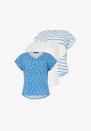 Vrouw met krullend haar, gekleed in een wit T-shirt met korte mouwen, opgerolde blauwe spijkerbroek en witte sneakers, staand tegen een effen muur.