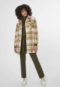 Geruite wollen jas in wit, geel en bruine tinten, met twee borstzakken en een relaxte pasvorm, gecombineerd met een olijfgroene ribbeltrui en broek.