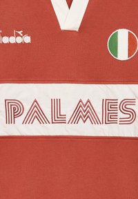 Maglia rossa con una fascia bianca che presenta il testo "PALMES" in modelli a strisce alternate. Include un emblema circolare della bandiera italiana.