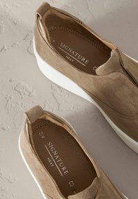 Chaussures slip-on en daim beige avec une semelle en caoutchouc blanc, dotées d'une poignée à l'arrière et d'un intérieur en cuir marron avec des marques imprimées.