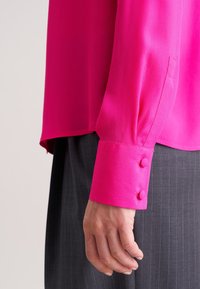 Lys pink langærmet bluse med glat tekstur og rund krave. Har knaplukning på ærmerne og er parret med et bukser med striber.