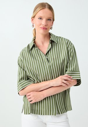 Woman with blonde hair wearing silver hoop earrings and a loose green shirt with white vertical stripes, posing with crossed arms.