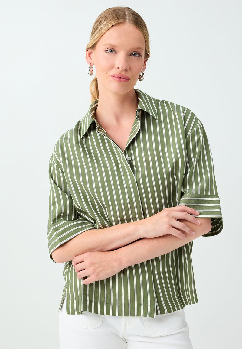 Femme aux cheveux blonds portant des boucles d'oreilles argentées en forme de créoles et une chemise verte lâche à rayures verticales blanches, posant les bras croisés.