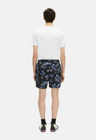 Schwarze Blumenshorts mit blauen und lila Mustern, kombiniert mit einem schlichten weißen T-Shirt. Das Model steht mit dem Rücken zum Betrachter und trägt Sneakers.