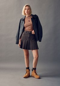 Camiseta marrón de manga larga con textura acanalada, falda plisada de denim negro y botas marrones con cordones y detalles en negro. La modelo está de pie frente a un fondo gris.