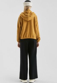 Senfgelbe Kapuzensweatshirt mit lockerer Passform, das aus strukturiertem Stoff gefertigt ist und einen elastischen Saum hat, kombiniert mit schwarzen weit geschnittenen Hosen.