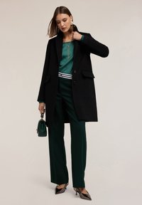 Cappotto di lana nero con vestibilità sartoriale, blusa di raso verde, pantaloni verdi e tacchi neri lucidi. Accessoriato con una piccola borsa a tracolla verde.