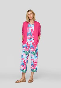 Helles rosa Strickjacke über einem blümchenmusterierten Top und weit geschnittenen Hosen. Der Stoff zeigt lebendige blaue, rosa und grüne Blätter auf einem hellen Hintergrund.
