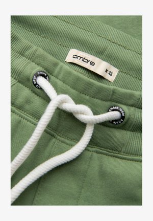 Pantaloni corti in cotone verde con texture a costine, dotati di un laccetto bianco e occhiello nero. L'etichetta riporta "ombre", taglia M, EUR 46.