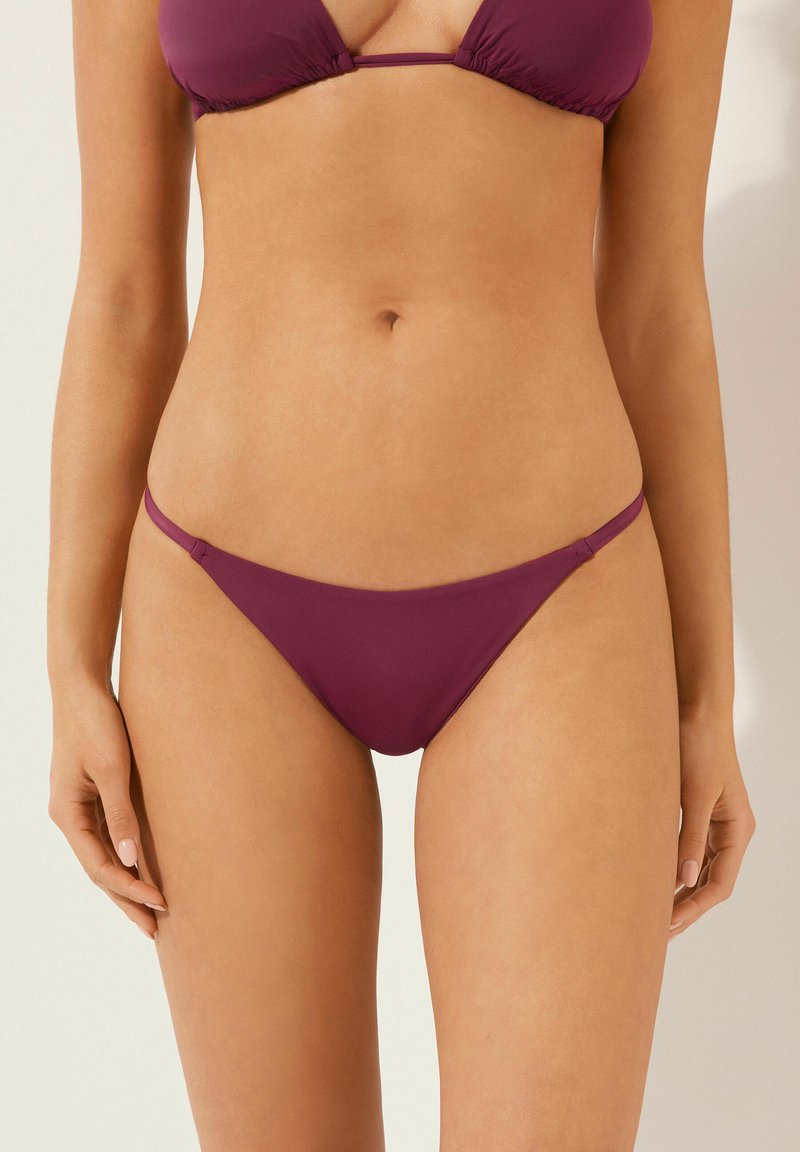 Bikinový set v tmavě burgundské barvě, obsahující triangelový vršek a spodní část s minimálním zakrytím a tenkými popruhy. Hladká látka bez viditelných vzorů.