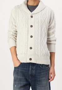 Krämfärgad stickad kofta med sjalkrage, flätstickade mönster och fem bruna knappar. Ribbade muddar och nederkant. Bärs över blåjeans.