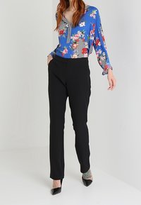 Donna che indossa pantaloni neri, décolleté neri a punta e una blusa floreale blu con accenti a righe, in piedi contro uno sfondo semplice.