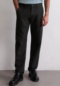 Schwarze Hosen aus glattem Stoff im geraden Schnitt, mit einem Knopfverschluss und seitlichen Taschen. Getragen mit schwarzen Schnürschuhen.