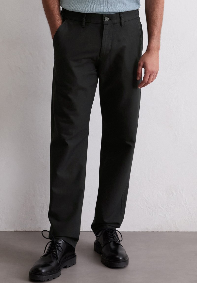 Pantaloni neri realizzati in tessuto liscio con design a gamba dritta, dotati di chiusura con bottone e tasche laterali. Indossati con scarpe nere stringate.
